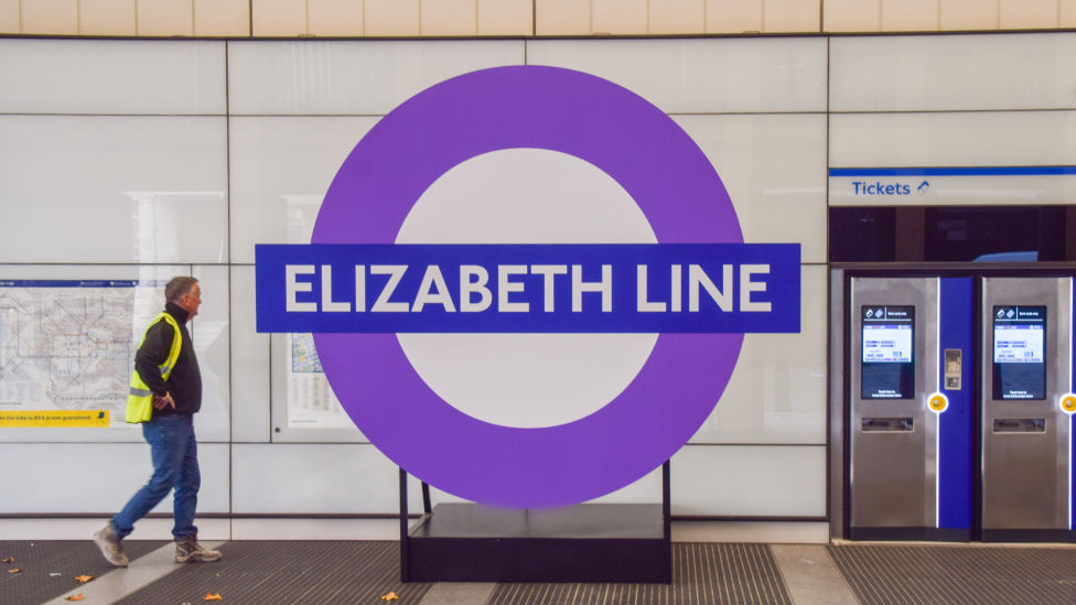 A man wearing a high viz jacket walks past an Elizabeth sign on a station platform