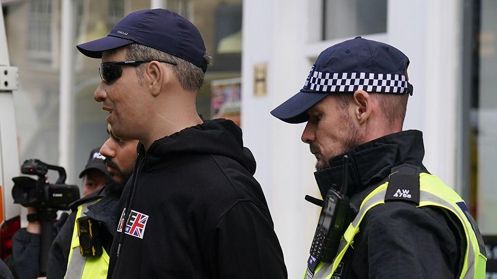 Police police offices in hi-vis vest detaining a protester wearing a rubber facemask, with the join visible on his neck, during the anti-immigration demonstration in Newcastle