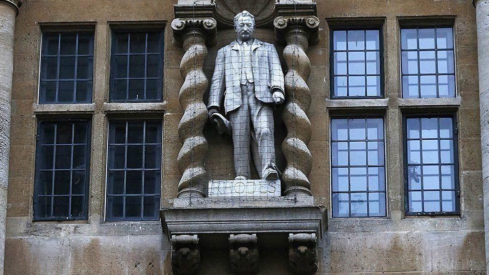 A statue of Cecil Rhodes, which stands on the outside of Oriel College. Rhodes is in a suit and waistcoat, carries a hat, and has hair and a moustache. The statue is behind a mesh.