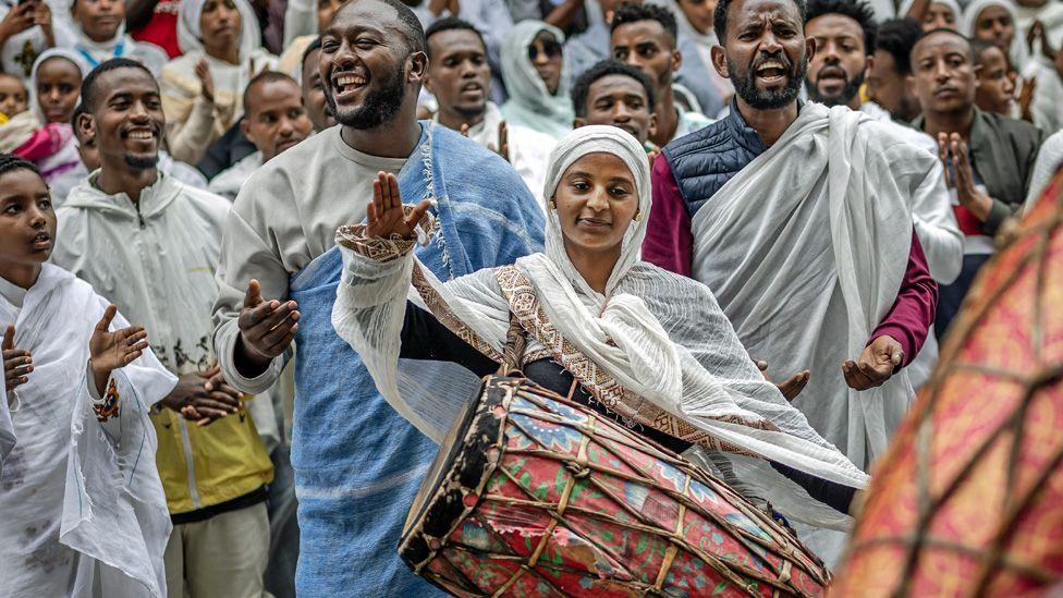 A crowd, including a woman in a traditional white shawl who plays a huge painted drum, sing New Year songs at Entoto St Raguel Church in Addis Ababa in Ethiopia - Thursday 11 September 2025.