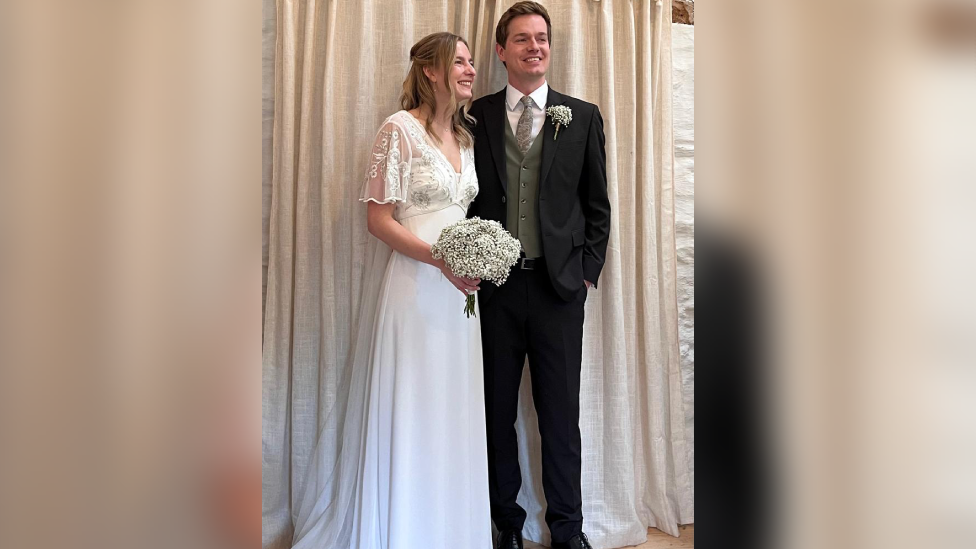 Rebecca wearing a floor-length white wedding dress and holding a bouquet of flowers, stood next to George who is wearing a three-piece suit with green tie 