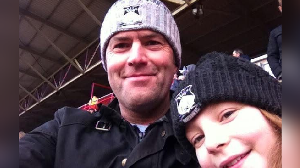 A close up selfie of Lili and her dad smiling whilst watching football at a Wrexham match. Lili, who is on the right, is wearing a black Wrexham bobble hat. Her dad, on the left, is wearing a black jacket and a grey Wrexham bobble hat.