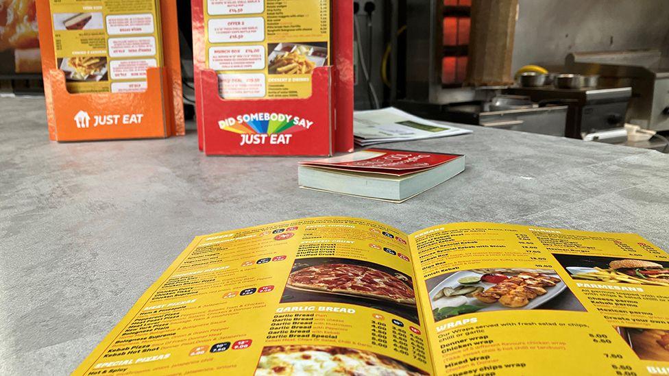 A yellow takeaway leaflet seen close up with more leaflets in red and orange stands behind. Further back there is a doner kebab in front of an upright grill.