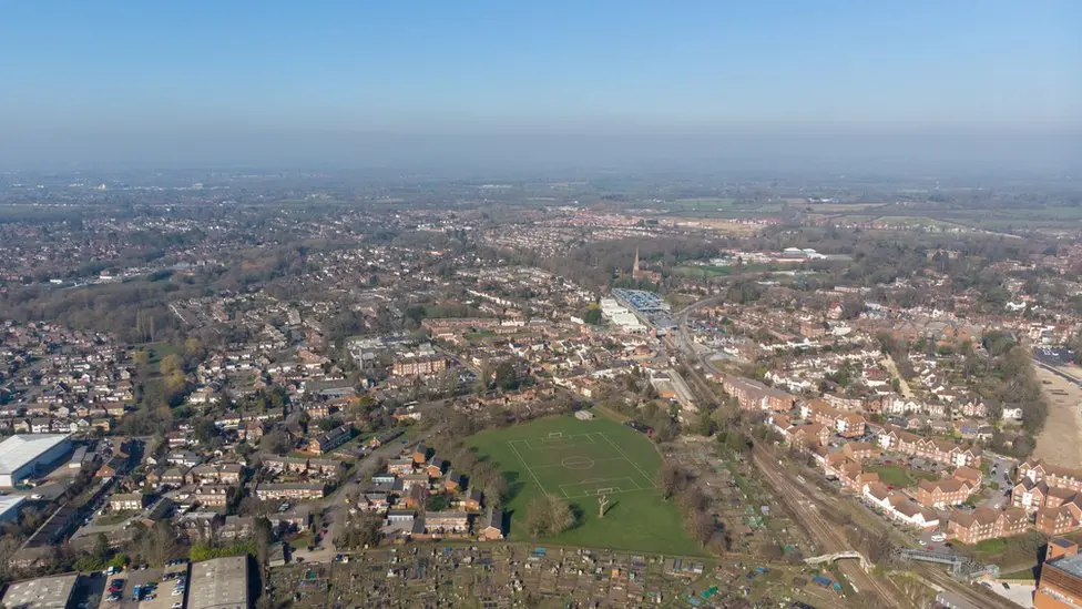 A bird's eye view over Wokingham, including a football pitch, a train line, homes, a factory to the left of the picture, and a church with a steeple in the middle of it