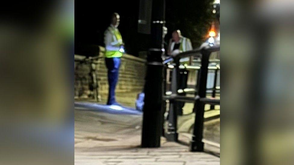 A blurry image showing two men wearing high visibility jackets on the Clifton Suspension Bridge. They are both shining large torches on a silver suitcase which contained some of Paul's body parts.