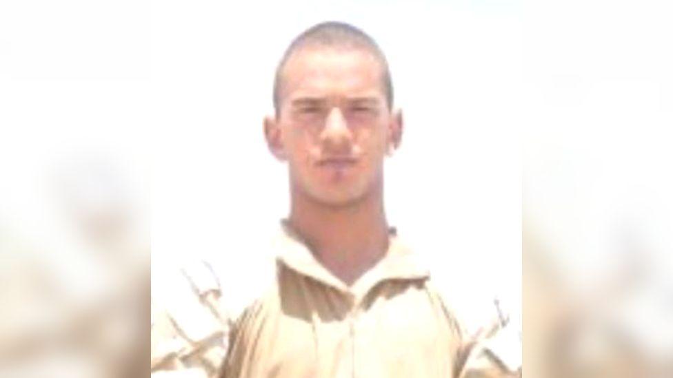 A blurry image of Mark Barrs, a man with short shaved hair, in beige-coloured military uniform