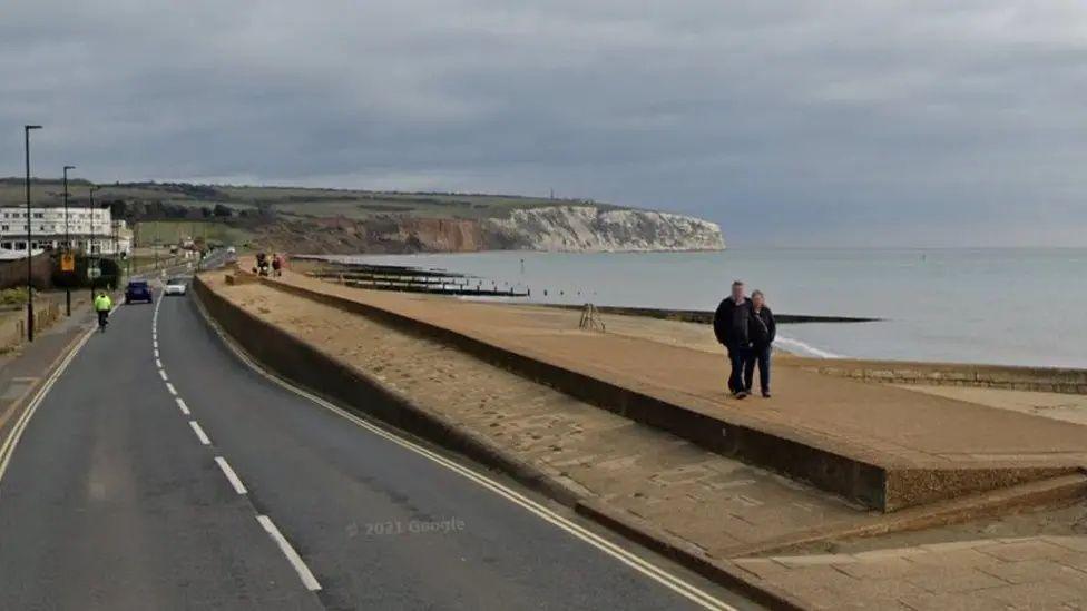 A Google image showing two people walking along the sea defences between a beach and a road with cliffs in the background. 