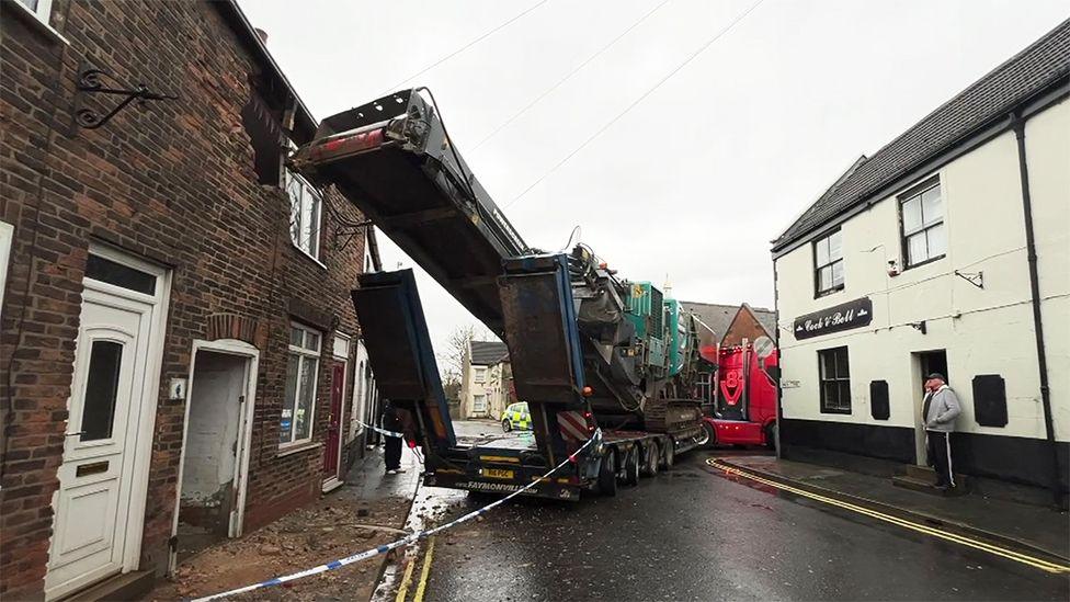 Another view of the crash showing the damage to the top floor of the house with debris on the street and blue and white police tape cordon around the vehicle with a person looking on from the door of the pub opposite