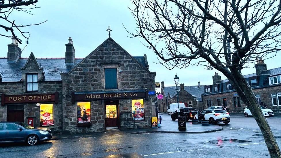A Post Office and a shop in Tarves town centre, with cars parked on the road, on a rainy day.