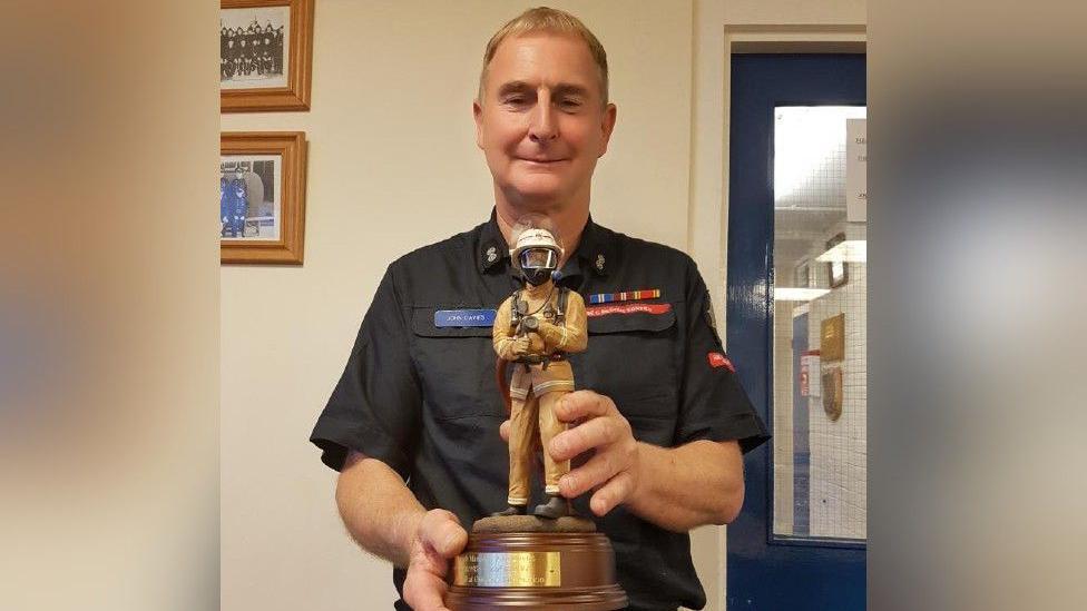 John Davies has short light brown hair and is wearing a dark shirt with a Shropshire Fire and Rescue service patch on its breast. He is holding an award trophy in the shape of a firefighter in full uniform with a helmet 