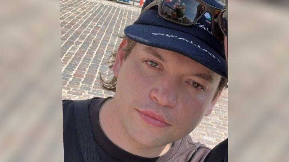 Luis Piovesana wearing a navy cap with brown sunglasses over them. He has short dark hair tucked behind his ears and is wearing a black T-shirt. He is clean shaven and is looking straight at the camera with a blank expression.