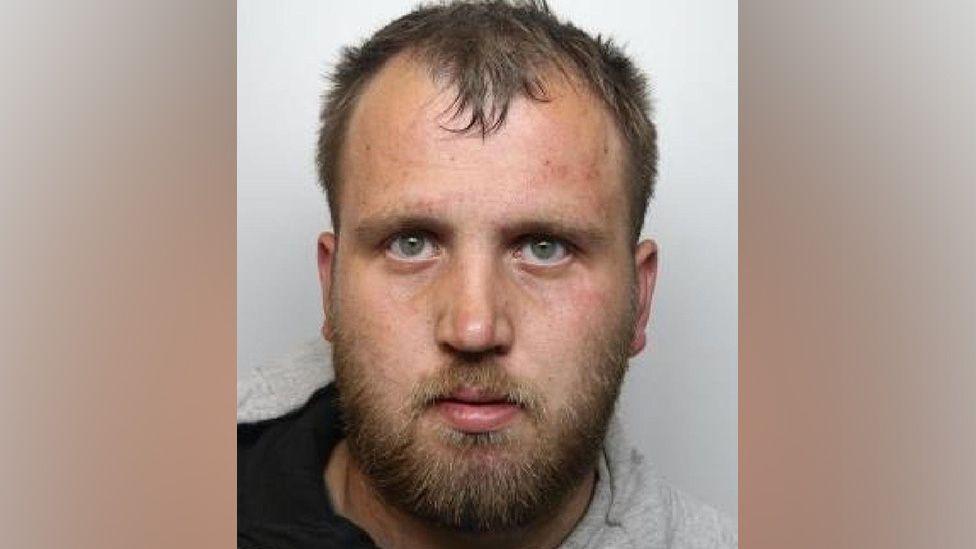 A mugshot of Joshua Simpson who has a brown beard, green or blue eyes and a receding hairline