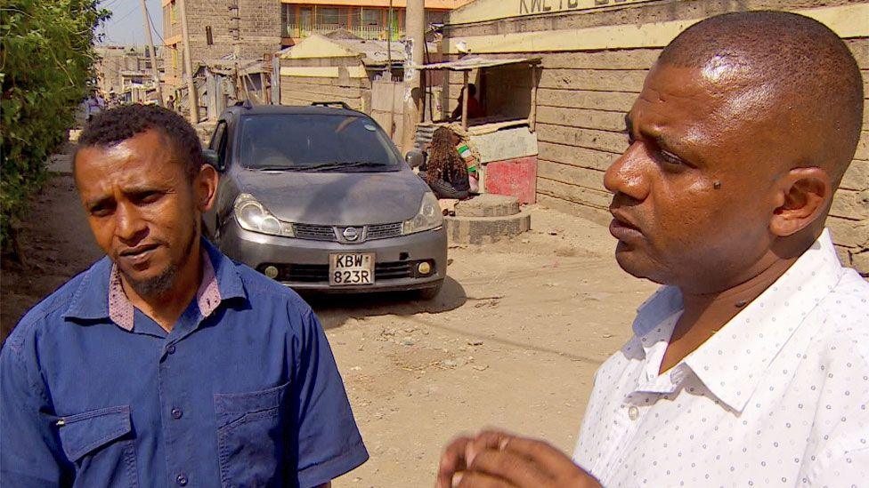 Aslam Longton in a blue shirt and Jamil Longton and a white shirt with a dot pattern talking as they stand on a dirt road in the town of Kitengela. A silver car can be seen behind them.