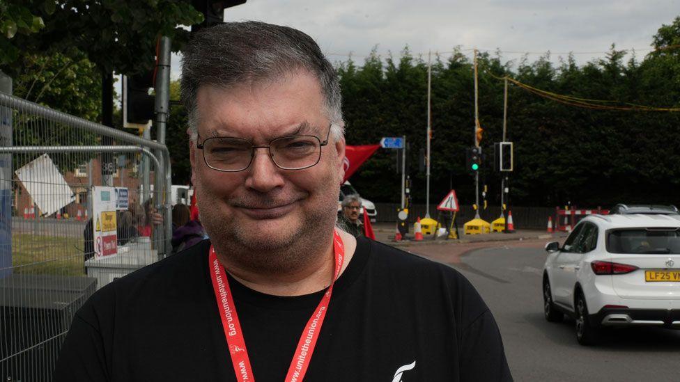 Paul Hardingham outside Addenbrooke's Hospital in Cambridge. Has greying black hair and is wearing glasses and a black T-shirt. Behind him can be seen roadworks and a roundabout.