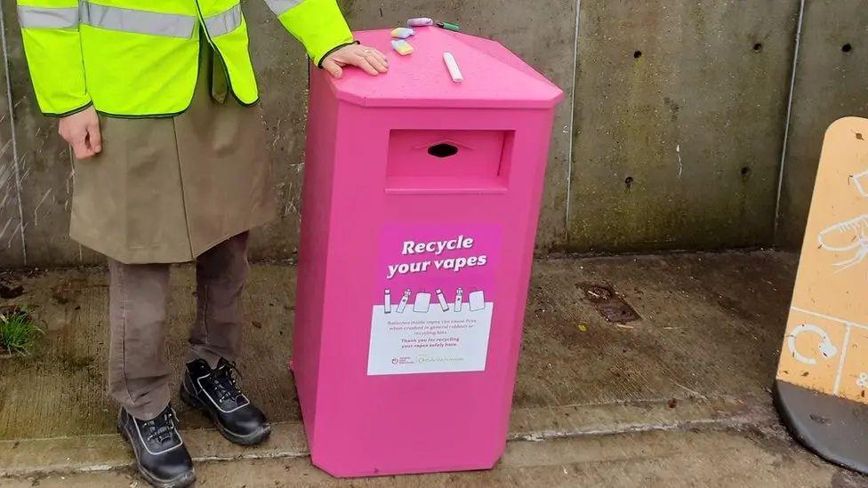 A person wearing beige trousers, black trainers and a high-vis jacket standing next to a bright pink bin with a label that reads: "Recycle your vapes".