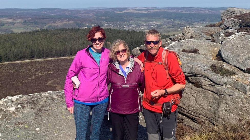 Naomi, Janice and Ray Craven, arm in arm, posing for a photograph next to some boulders on a fell top, with woods and countryside in view behind them.