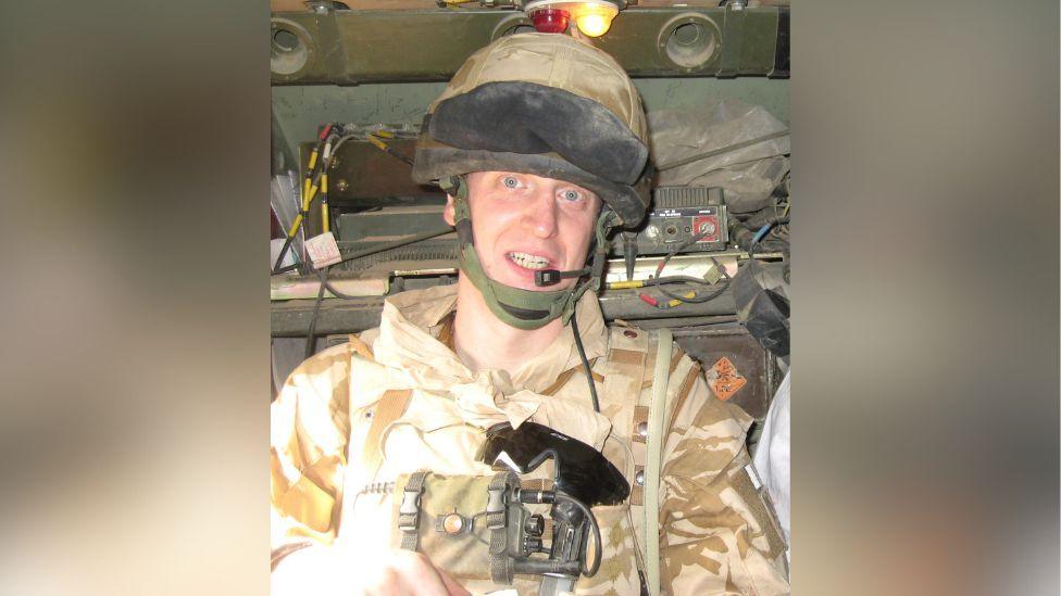 Rob Shenton is pictured inside a military vehicle in Afghanistan. He is wearing desert camouflage including a helment and radio mouthpiece. Sunglasses are attached to his chest and military equipment is pictured behind him