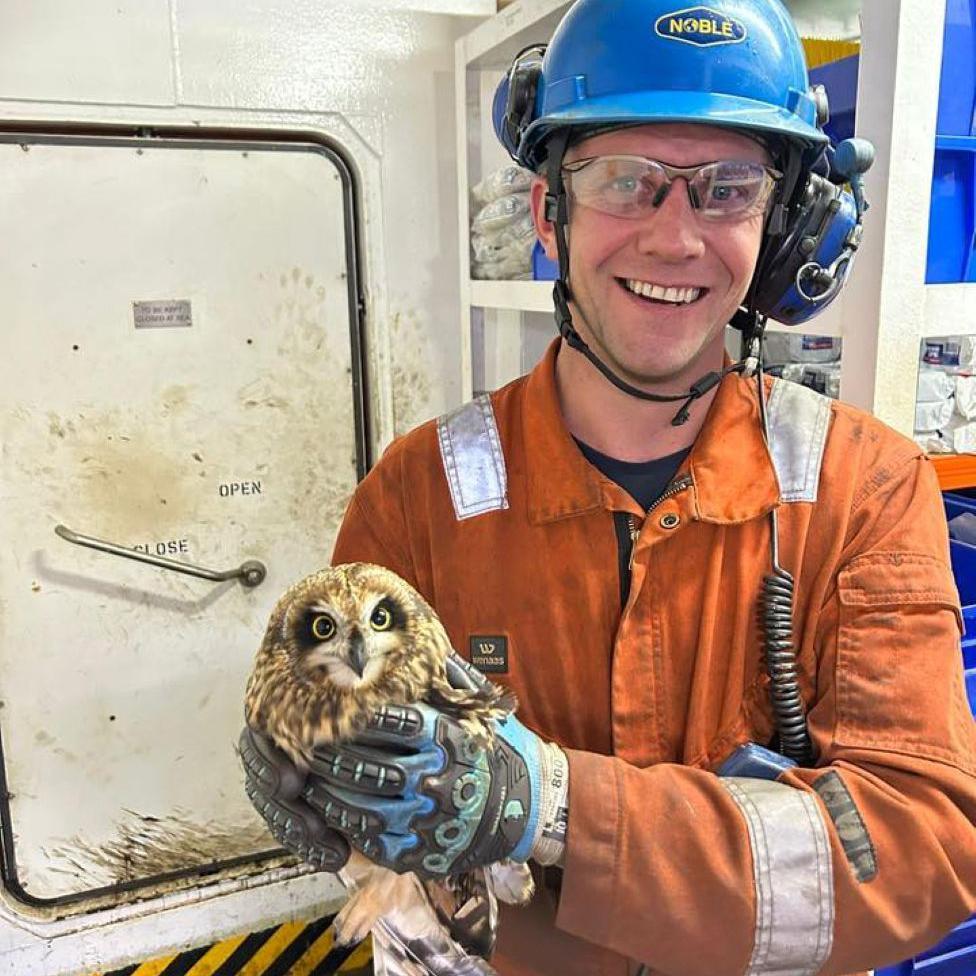 Offshore worker Sam Crowe in orange overalls, smiling at camera, and holding an owl.