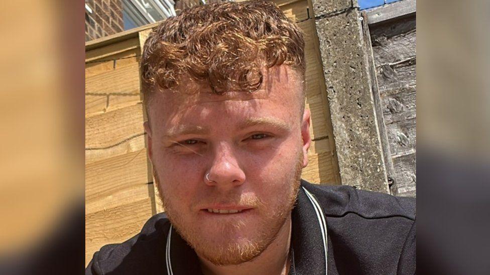 Levi Kent, with ginger curly hair and a short ginger beard, and wearing a black T-shirt, pictured at close range against a brown fence