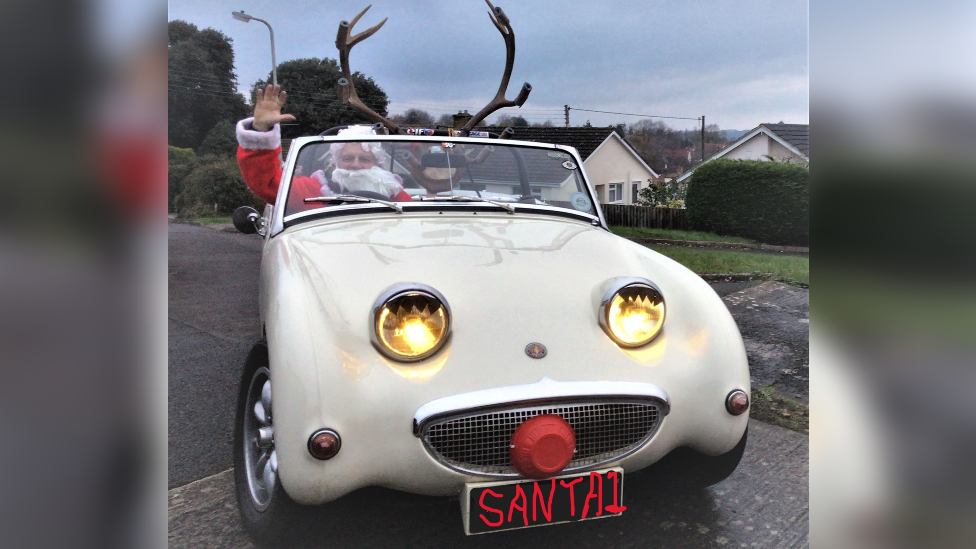 A 1979 MG Midget Frogeye Sprite replica with antlers attached to the front, and Robin in the driver's seat dressed as Santa Claus