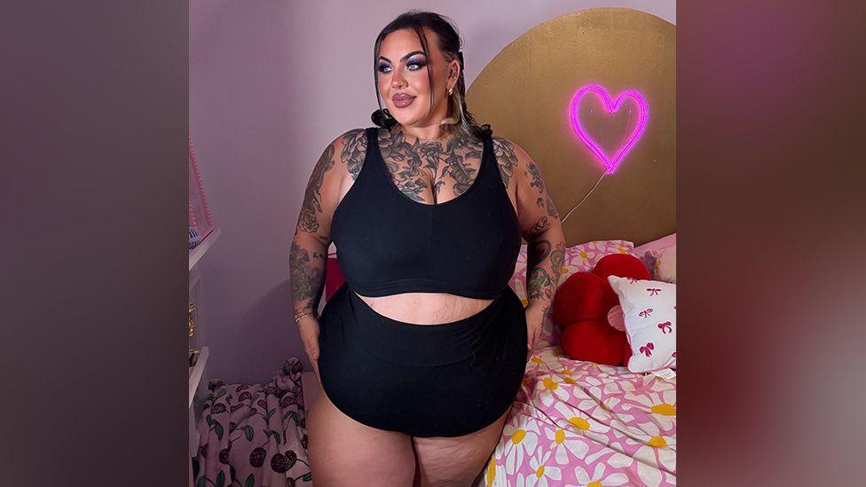 Sophie who is a UK size 24 to 26 model wearing a black sports bra and cycling shorts and glamorous make up, posing in a pink bedroom in an Instagram-style shot