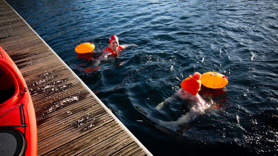 James treads water at the half-way point of his swim. Only his head is above water and he is wearing an orange bobble hat. A swimming buoy is attached to him. There is another swimmer in the water next to him.