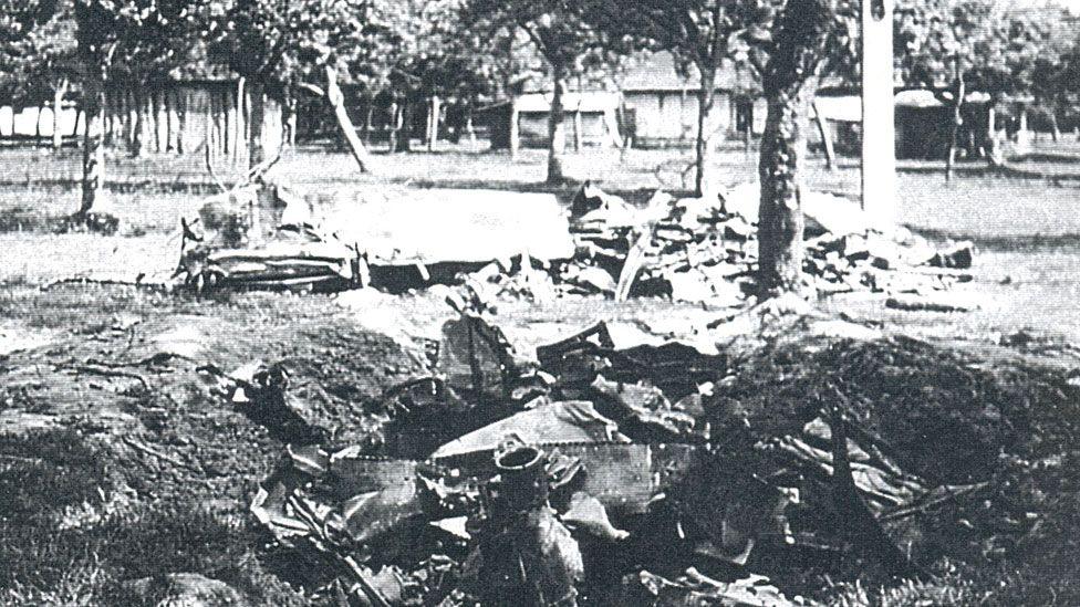 A black and white image showing scattered debris belonging to a crashed WW2 fighter plane. Debris is buried in a crater. Beyond it are trees and farm buildings. 