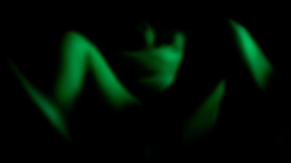 Artwork created by Phoebe Wingrove to depict the experience of traumatic birth, it is an abstract photograph where you can just make out the raised knees of a woman sitting back and viewed from her feet. Her body has taken on a green colour and the rest of the scene is in black.