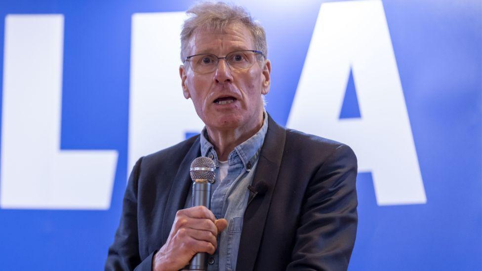 Kenny MacAskill, a grey-haired bespectacled man, speaks at an event while holding a silver microphone in front of his chest. He is wearing a dark jacket over a denim shirt. A blue Alba sign is in the background. 