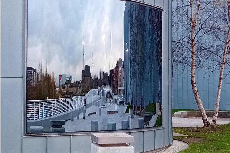 There is a warped-looking bridge in the refection and some bollards with some buildings in the background of the reflection. There is a tree at the side of the building. 