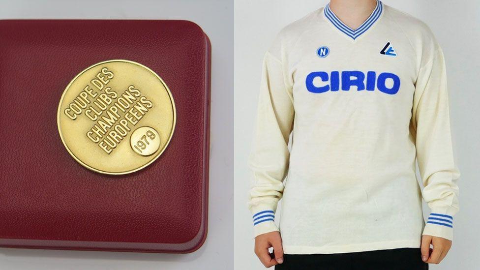 There are two seperate images here. On the left is a photograph of Francis' European Cup medal. It's golden and has "COUPE DES CLUBS CHAMPIONS EUROPEENS - 1979" engraved on it. On the right hand side is a photograph of the match worn Maradona shirt that sold for £20,000. It's an off white colour with a striped blue v-neck collar and striped blue sleeve cuffs. There is a sponsor in the middle which says "CIRO" in blue.