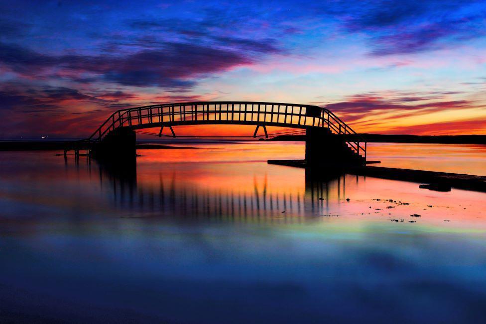 Sunrise sky of purple, blue, orange and yellow, reflected in water, with a bridge across the water.