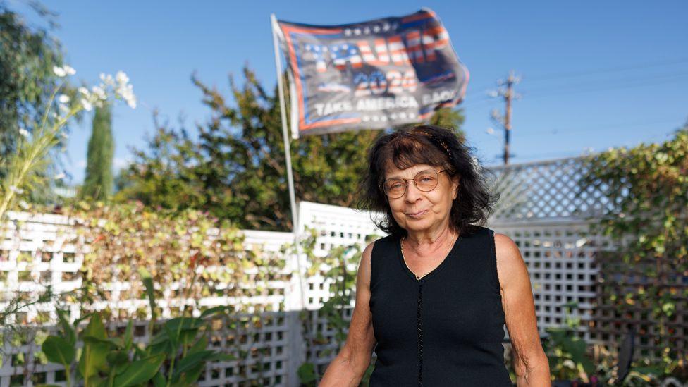 June in her garden, looking at the camera smiling, in front of a white fence with plants, a blue sky and a flag printed with the words Trump 2024.  She is wearing a black cropped top and round wireframe glasses.