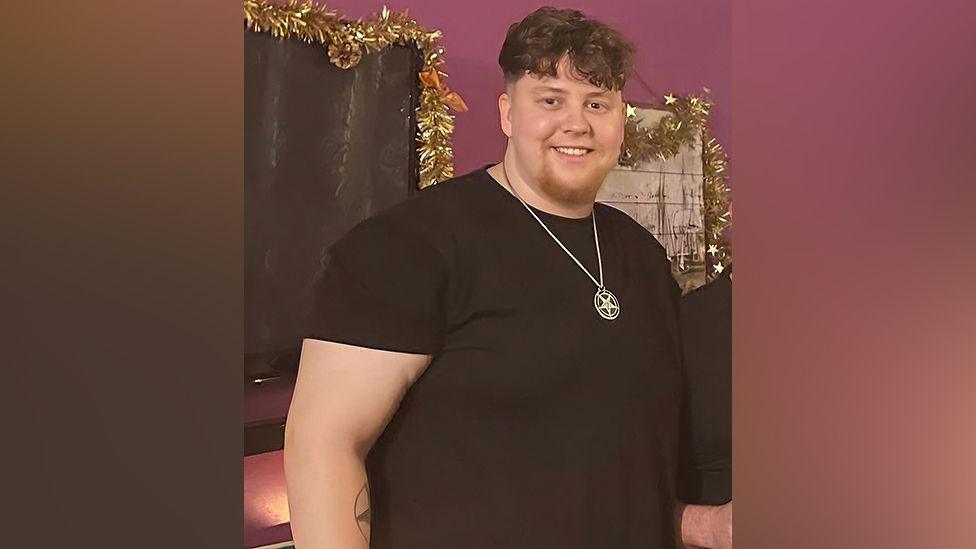 Jake Baxter is wearing a black top and silver medallion around his neck. He is staring at the camera and smiling. He is pictured at his heaviest, weighing 25 stone (159kg).