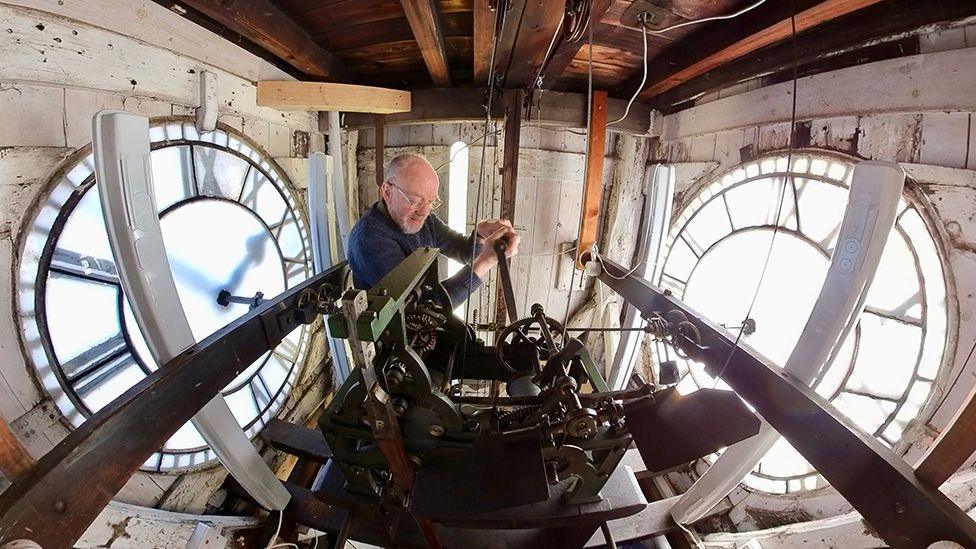 Graham Tebbs inside the clock tower and behind the clock workings, using a long winding handle. On either side of him are the large, white clock faces with Roman numerals. Mr Tebbs has white receding hair, a white beard and glasses.