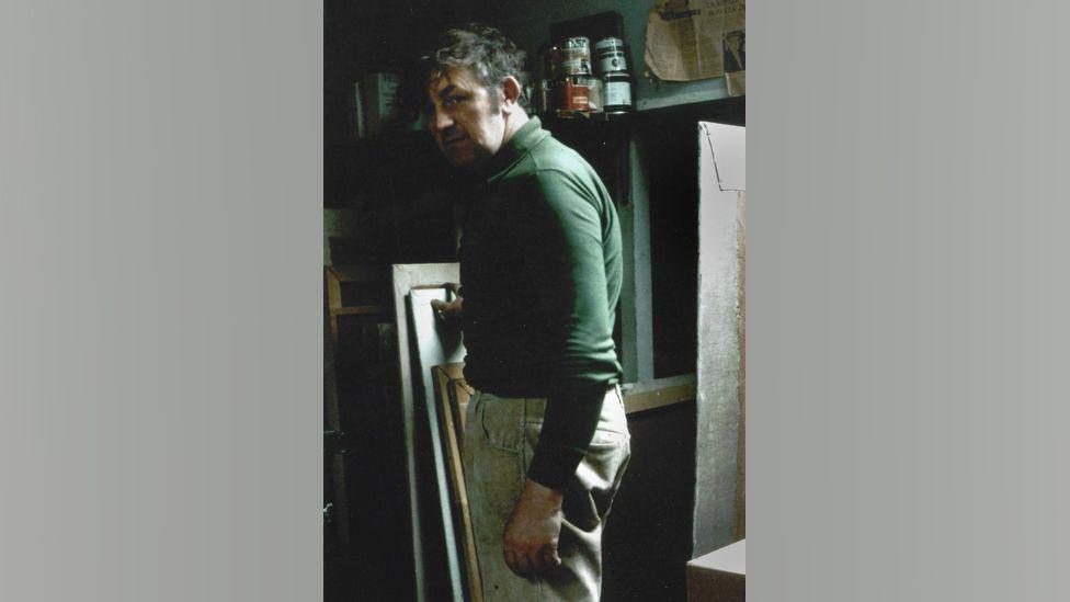 A young Tom McGuinness in his loft space studio in Bishop Auckland. He is standing side on to the camera and is wearing a green top. He is standing in front of a number of canvases and tins of paints.