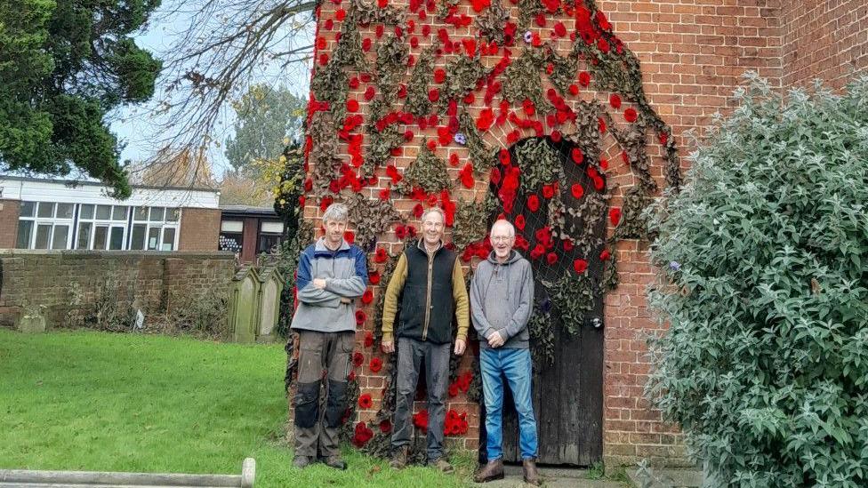 Thousands of red and purple knitted poppy flowers are attached to dark green netting, which is draped down the side of a red brick church tower. Three men are standing in front of the cascade, which falls over a brown wooden door at the bottom