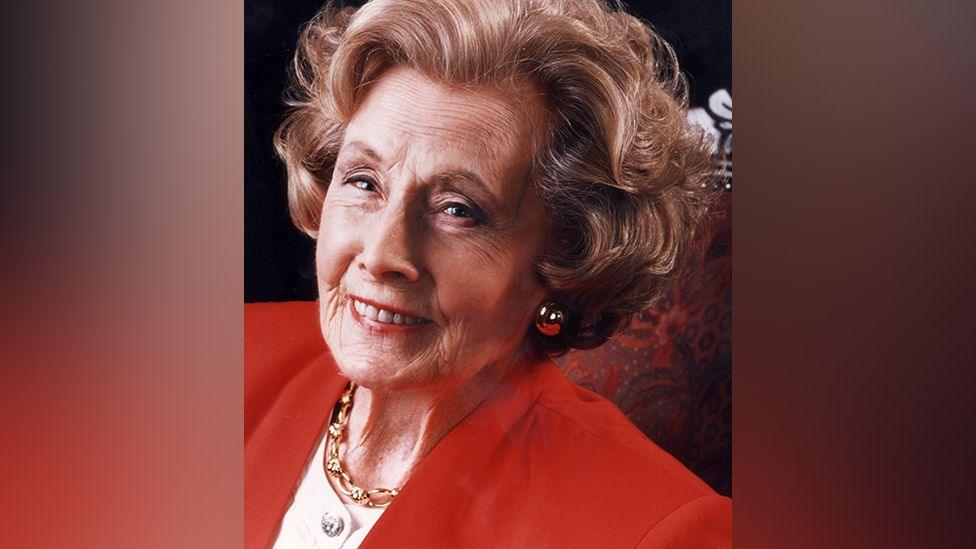 The late Baroness Barbara Castle smiling wearing a red jacket and cream top 