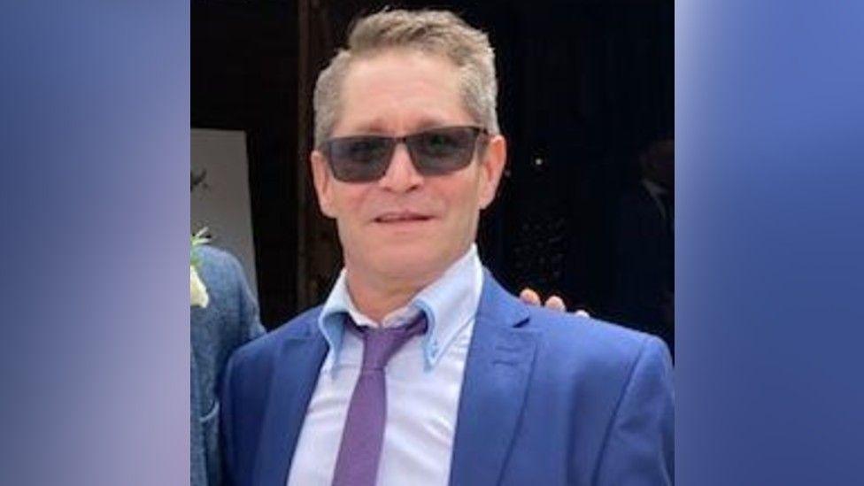 Robert Brown, a man who is standing outside. He is wearing a blue suit jacket, pale blue shirt and purple tie. He is also wearing a pair of sunglasses over his eyes and is looking directly at the camera and smiling.
