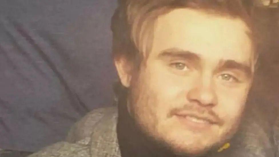 Jac Lewis smiles at the camera, he has long sandy hair and is wearing a grey blazer.