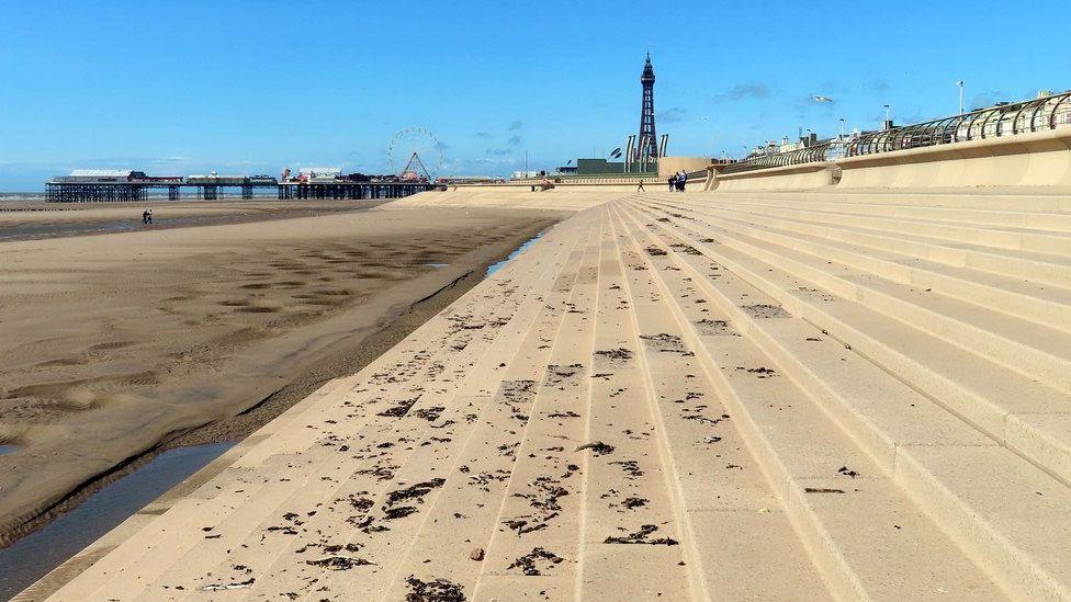 Steps leading on to Blackpool beach, with the tide out. Te tower and pier can be seen in the distance against a blue sky