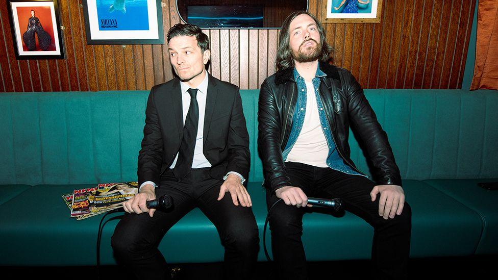 The two members of the band Benefits, Kingsley Hall and Robbie Major, sitting on a teal-coloured sofa in front of a dark, wood panelled wall, both holding microphones on their laps. Kingsley Hall is wearing a black suit, white shirt and black tie. Robbie Major is wearing black trousers and a black leather jacket over an open denim shirt and white t-shirt. 