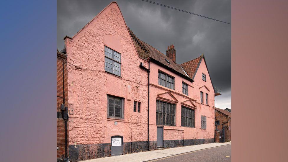 The Music House in Norwich. It is painted pink on the outside and has two gables, one on the right and one on the left. there are windows along the ground and first floor. There is also a window in the right gable and another can be seen in the roof