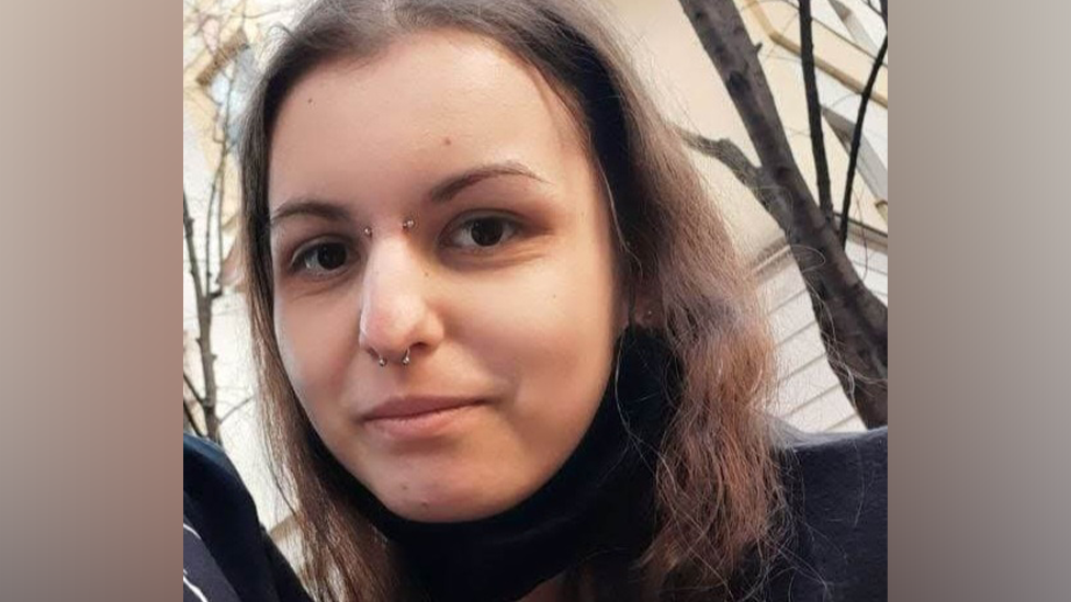 A woman with long brown hair, a brow piercing and a nose piercing, smiles at the camera. Trees without leaves are visible behind her.