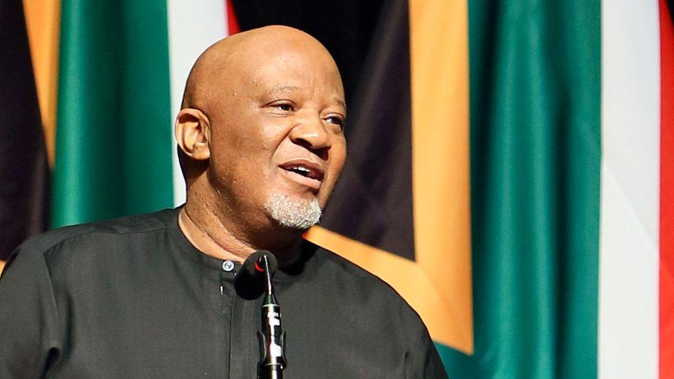 Mcebisi Jonas in a black collarless shirt speaks in front of microphone with the South African flags behind him.