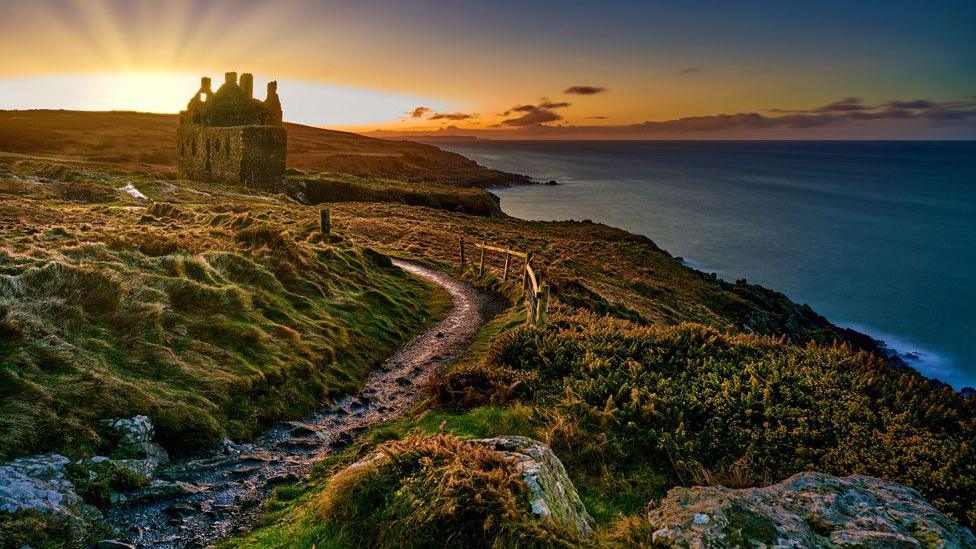 A bumpy path leads to a ruined castle on the Scottish coast with the sun peeping out above the horizon behind it.