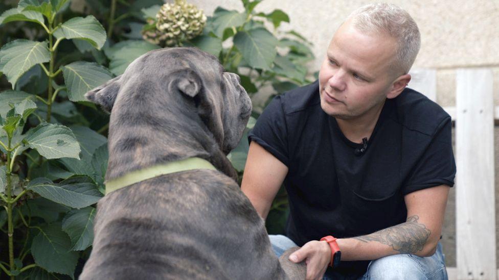 Francis sitting down in a black T-shirt, with short blond hair, holding the paws of a large dog