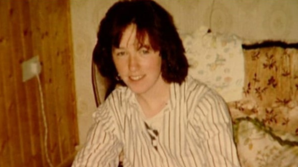 Jo Jo Dullard, wearing a white, stripey top, sat in front of a patterned chair and wood panelling and beige wallpaper