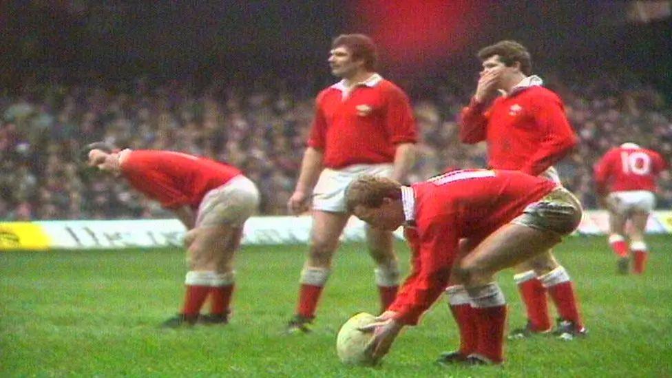 Paul Thorburn places a rugby ball on ground while Wales teammates in red jerseys look on