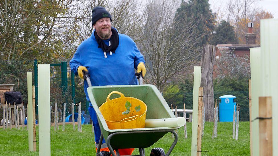 A man dressed in a blue shirt and wool hat pushes a wheelbarrow with a sapling in a yellow container through a row of recently planted trees